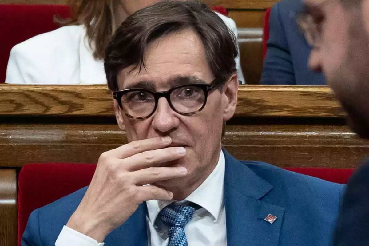 Hombre con gafas y traje azul sentado en un entorno formal, con la mano en la boca y expresión pensativa.