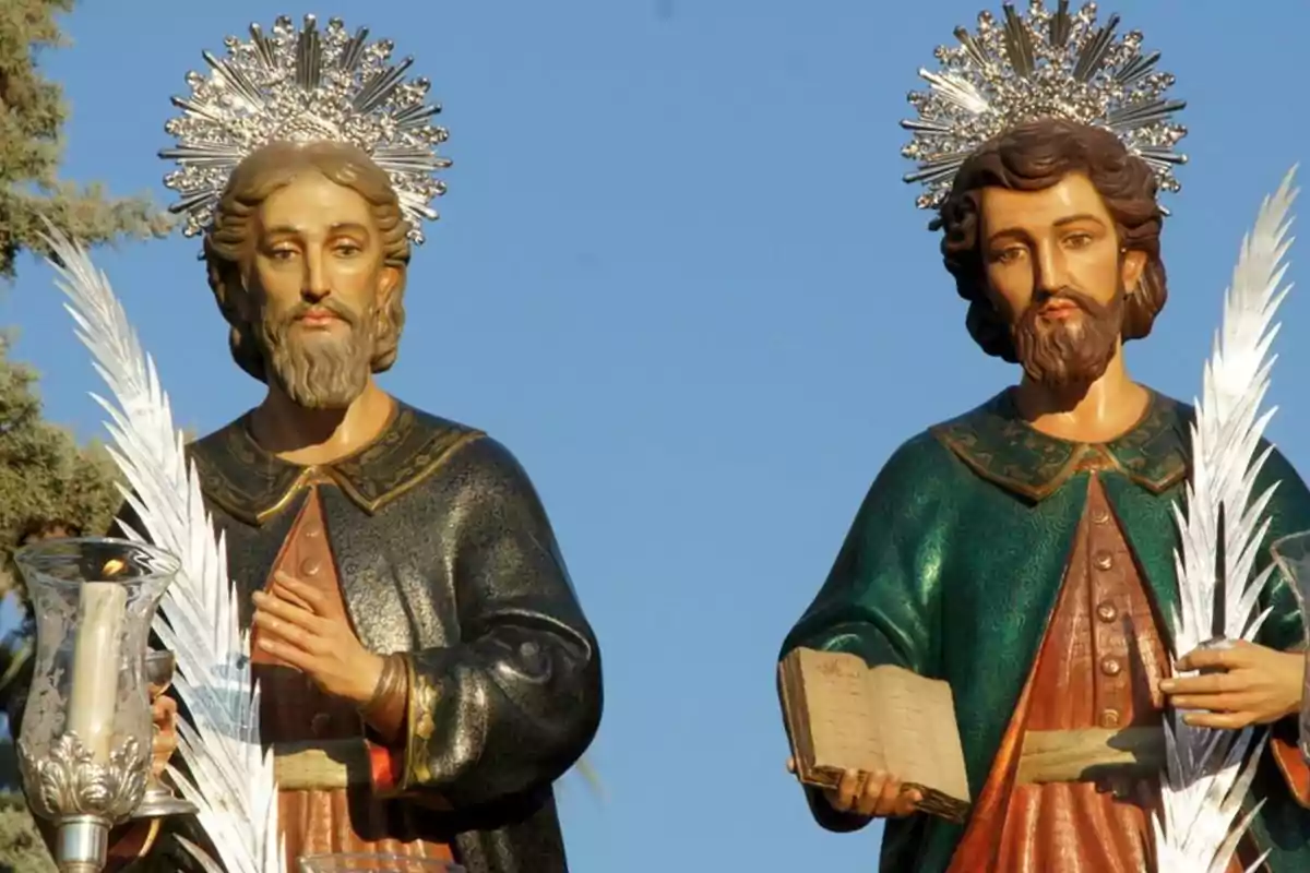 Dos estatuas religiosas con aureolas y palmas, una sosteniendo una vela y la otra un libro, con un fondo de cielo azul.