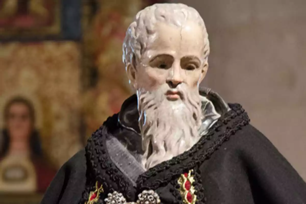 Escultura de un hombre con barba blanca y cabello rizado, vestido con una túnica negra decorada con detalles dorados y rojos, en un entorno con fondo borroso.