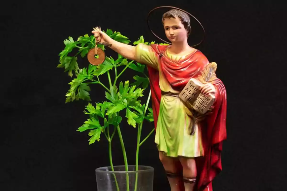 Una estatua de un santo con aureola sosteniendo una llave y un libro, junto a una planta verde en un vaso transparente, sobre un fondo negro.