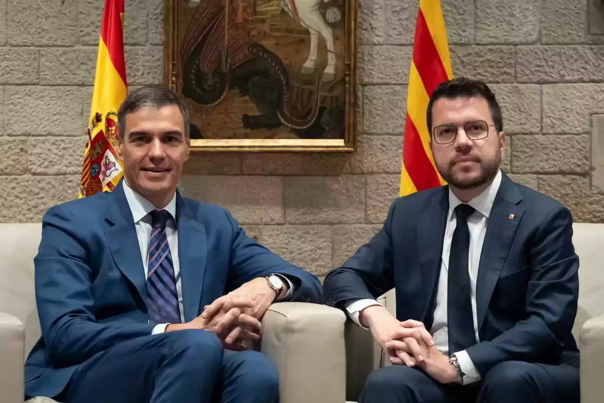 Dos hombres con trajes formales sentados en sillones, con banderas de España y Cataluña detrás de ellos y un cuadro en la pared.