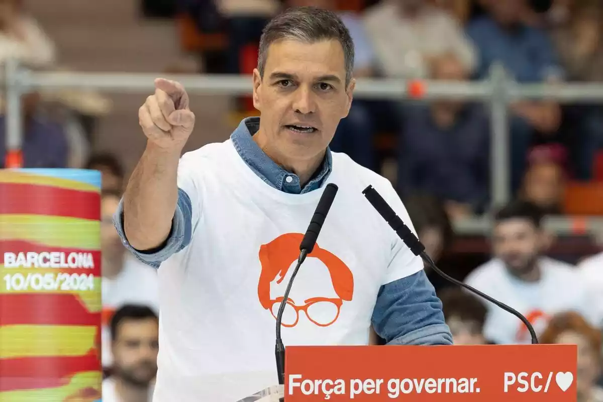 Pedro Sánchez en un mitin hablando desde un atril con una camiseta con el simbolo de la cara de salvador illa y señalando al frente