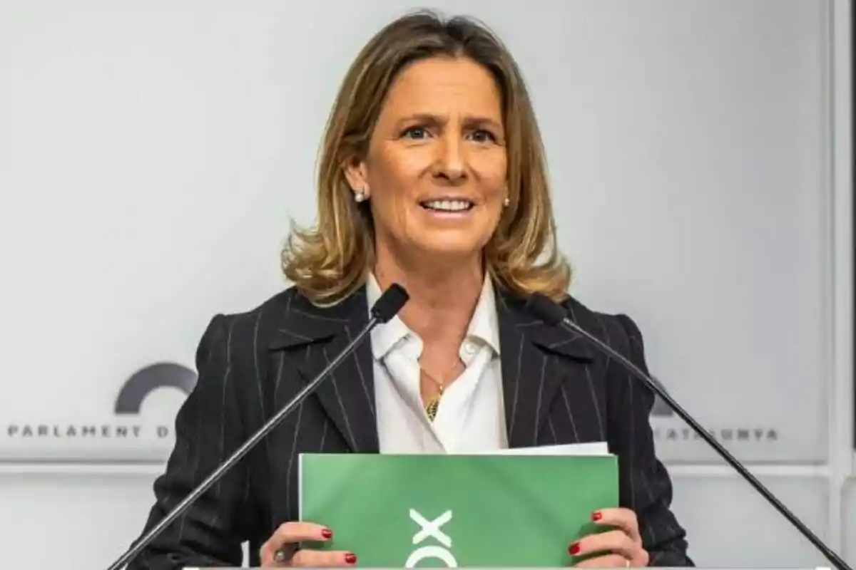 Mujer de cabello rubio y corto hablando en una conferencia de prensa con dos micrófonos y sosteniendo una carpeta verde