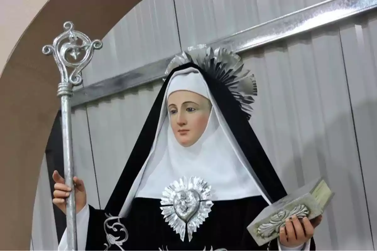 Estatua de una monja con hábito negro y blanco, sosteniendo un báculo plateado en una mano y un libro en la otra, con un corazón plateado en el pecho. Estatua de una monja con hábito negro y blanco, sosteniendo un báculo plateado en una mano y un libro en la otra, con un corazón plateado en el pecho.
