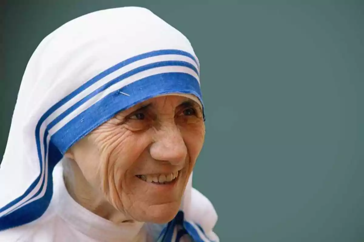 Una mujer mayor con un hábito blanco y azul sonríe frente a un fondo verde.