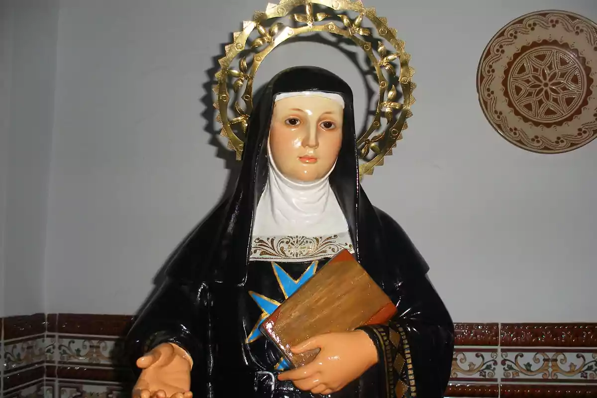 Una estatua religiosa de una figura femenina con hábito negro y un halo dorado sosteniendo un libro en una mano. Una estatua religiosa de una figura femenina con hábito negro y un halo dorado sosteniendo un libro en una mano.