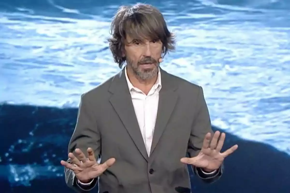 Santi Millán con barba y cabello largo, vestido con un traje gris, gesticula con las manos frente a un fondo de agua en Got Talent.