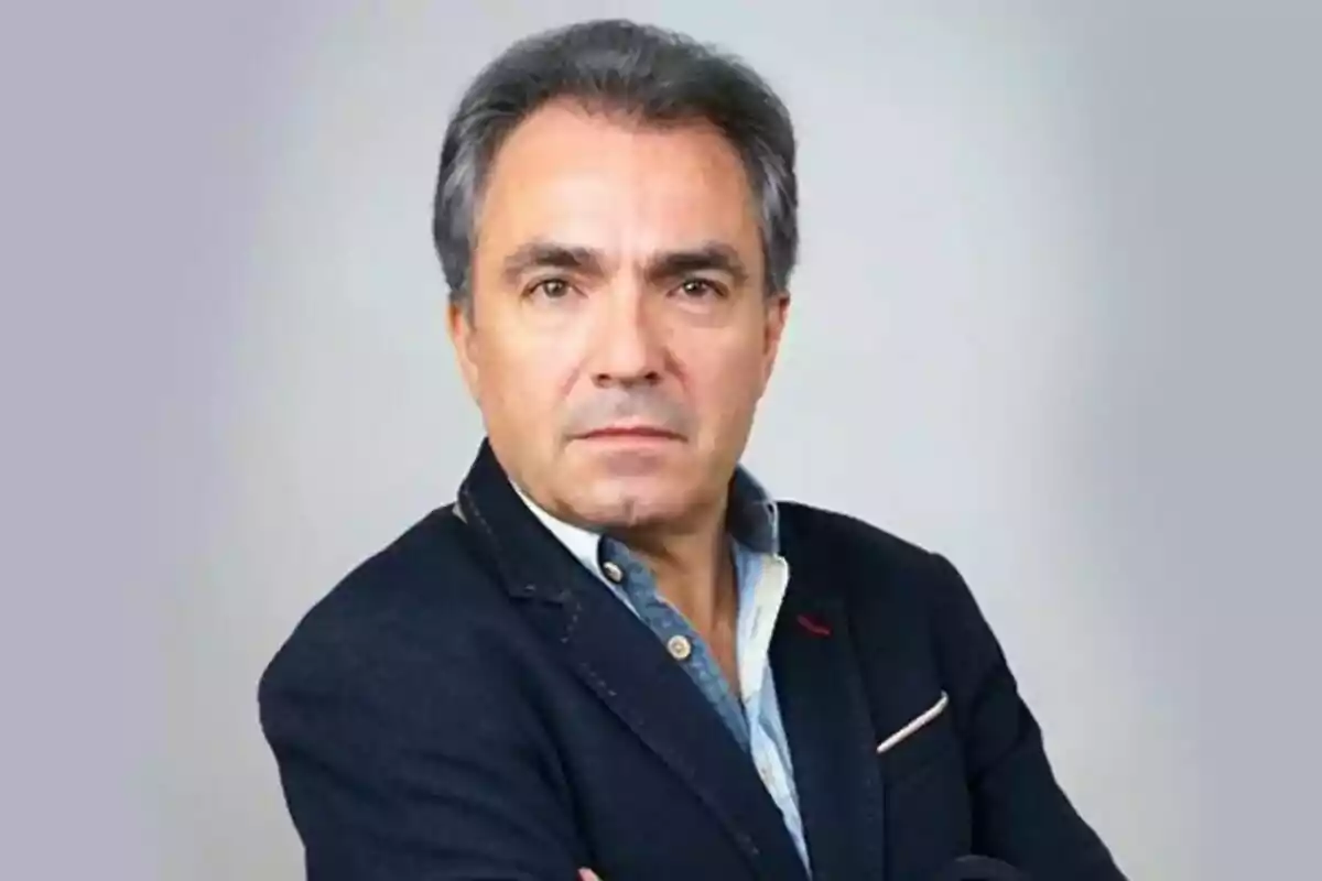 Hombre de mediana edad con cabello canoso y expresión seria, vestido con una chaqueta oscura y camisa clara, posando con los brazos cruzados frente a un fondo gris. Hombre de mediana edad con cabello canoso y expresión seria, vestido con una chaqueta oscura y camisa clara, posando con los brazos cruzados frente a un fondo gris.