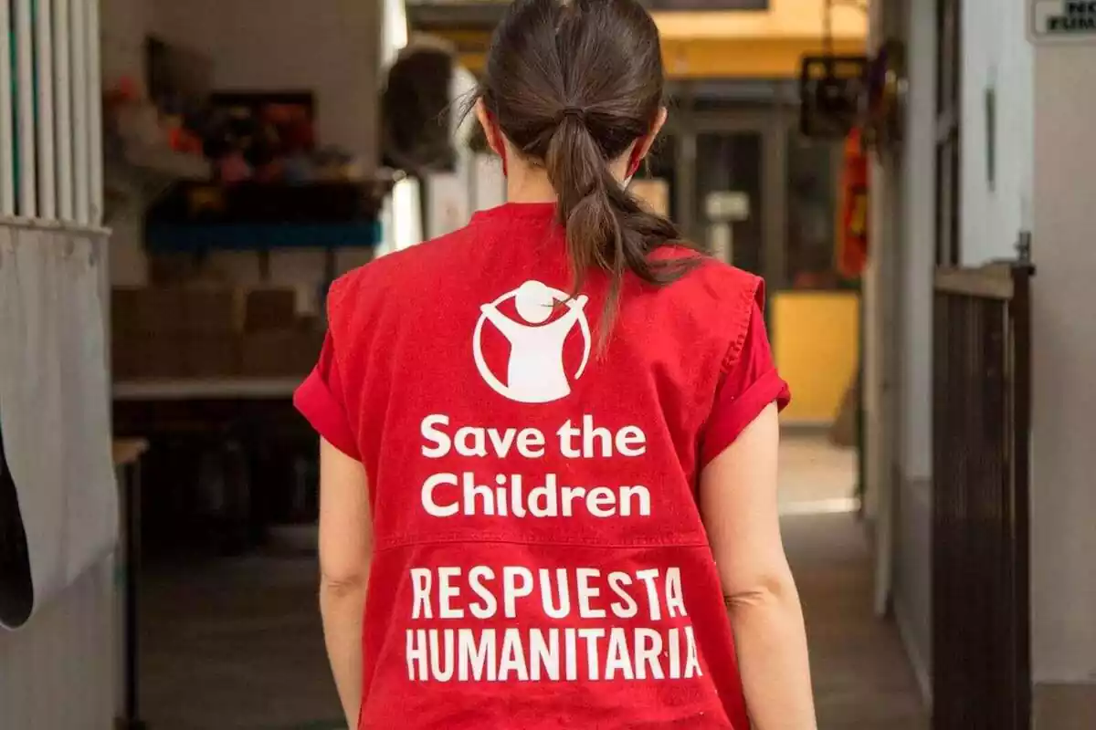 Plano medio de una voluntaria de la ONG Save the Children de espaldas y con un chaleco rojo con el logo de la ONG y la frase 'respuesta humanitaria'