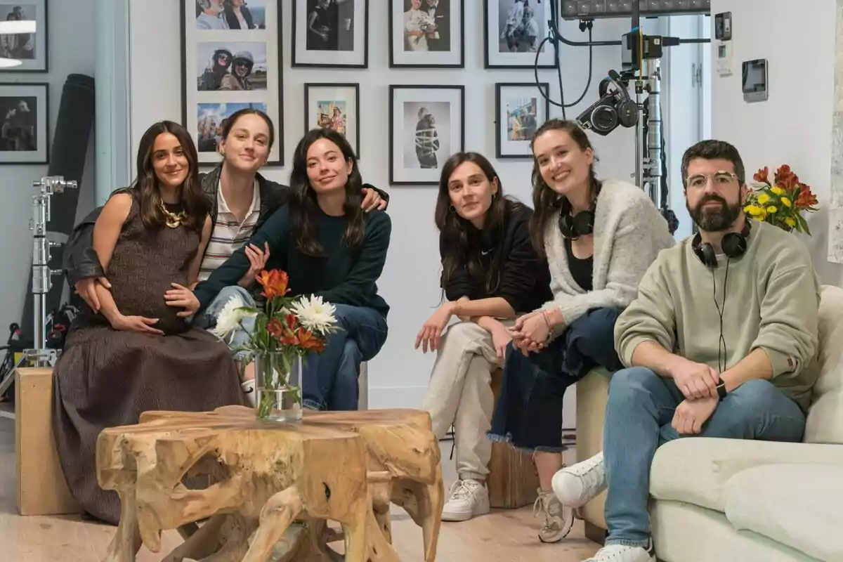 Anna Castillo, Macarena García, Laura Weissmahr, Victoria Martín, Sandra Romero y Nacho Pardo sentadas en una sala moderna con paredes decoradas con fotografías y equipo de grabación al fondo, todas sonríen y parecen relajadas, en el centro hay una mesa de madera con un florero durante el rodaje de Se tiene que morir mucha gente.