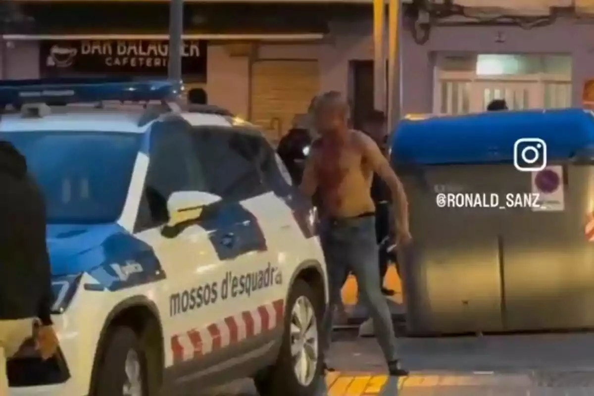 Un hombre sin camisa está de pie junto a un coche de policía de los Mossos d'Esquadra en una calle, con un contenedor de basura azul al fondo y una cafetería visible detrás.