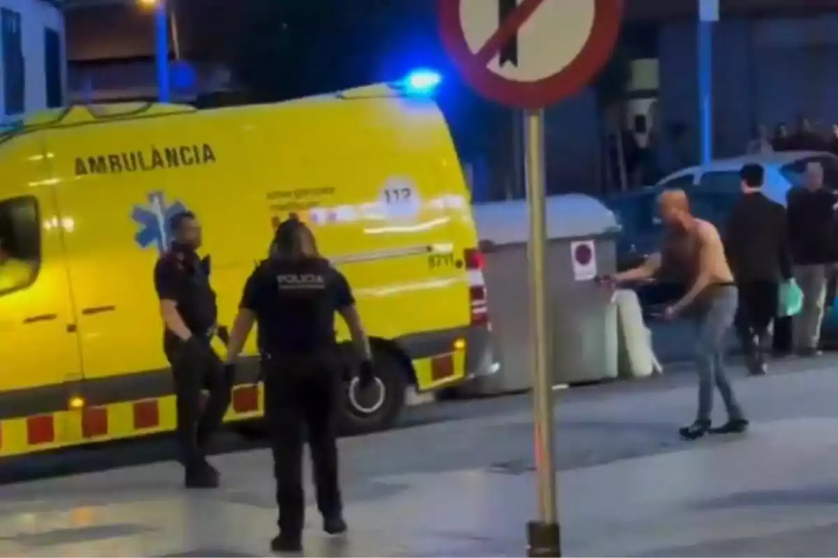 Una ambulancia amarilla está estacionada en la calle mientras dos policías interactúan con un hombre sin camisa cerca de un contenedor de basura. Una ambulancia amarilla está estacionada en la calle mientras dos policías interactúan con un hombre sin camisa cerca de un contenedor de basura.