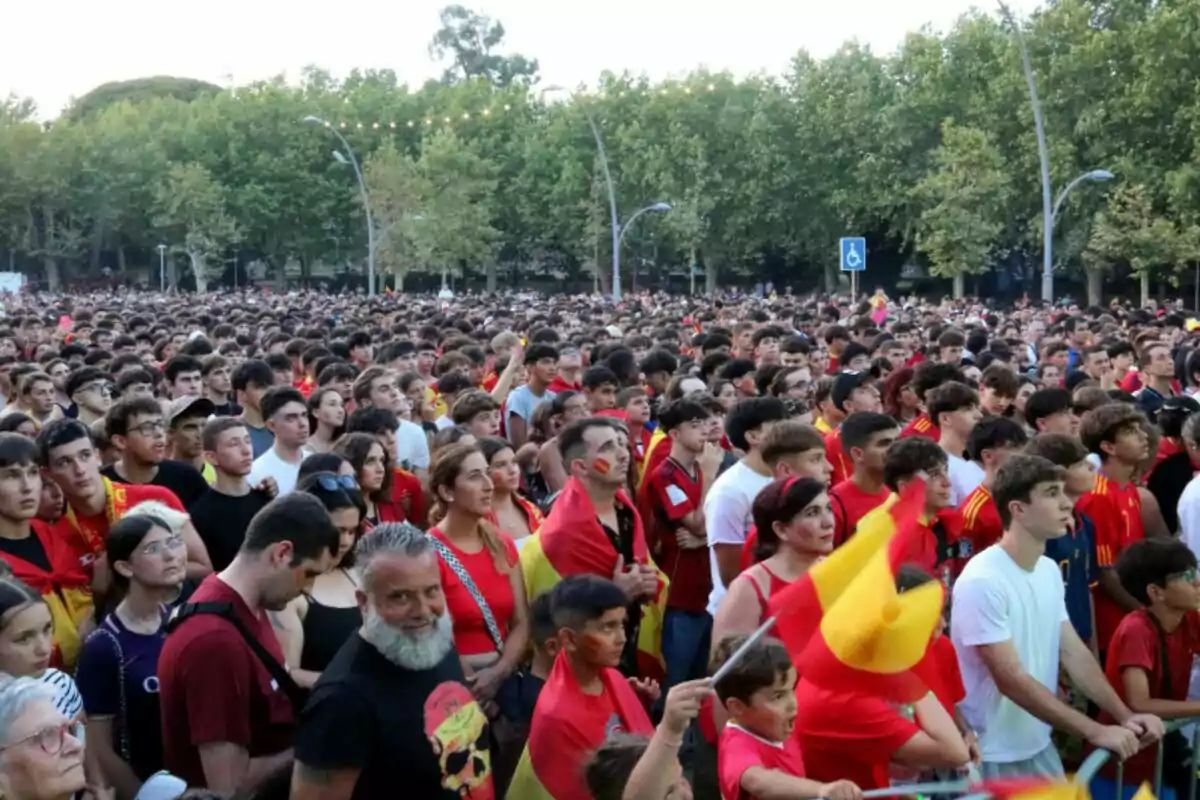 Una multitud de personas con camisetas y banderas de España se reúne en un parque con árboles al fondo.