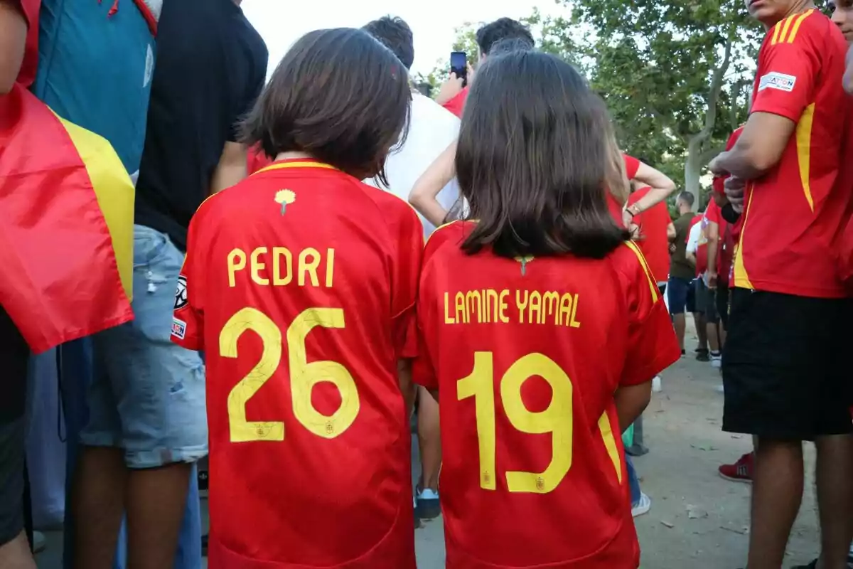 Dos niños de espaldas llevan camisetas rojas de la selección española de fútbol con los nombres "Pedri" y "Lamine Yamal" y los números 26 y 19, rodeados de más personas con camisetas similares.