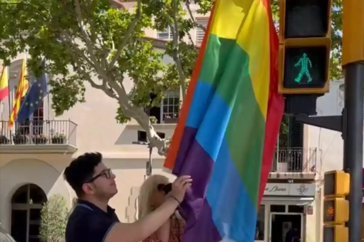 Una persona sosteniendo una bandera del orgullo LGBT+ junto a un semáforo en verde para peatones en una calle con árboles y edificios de fondo.