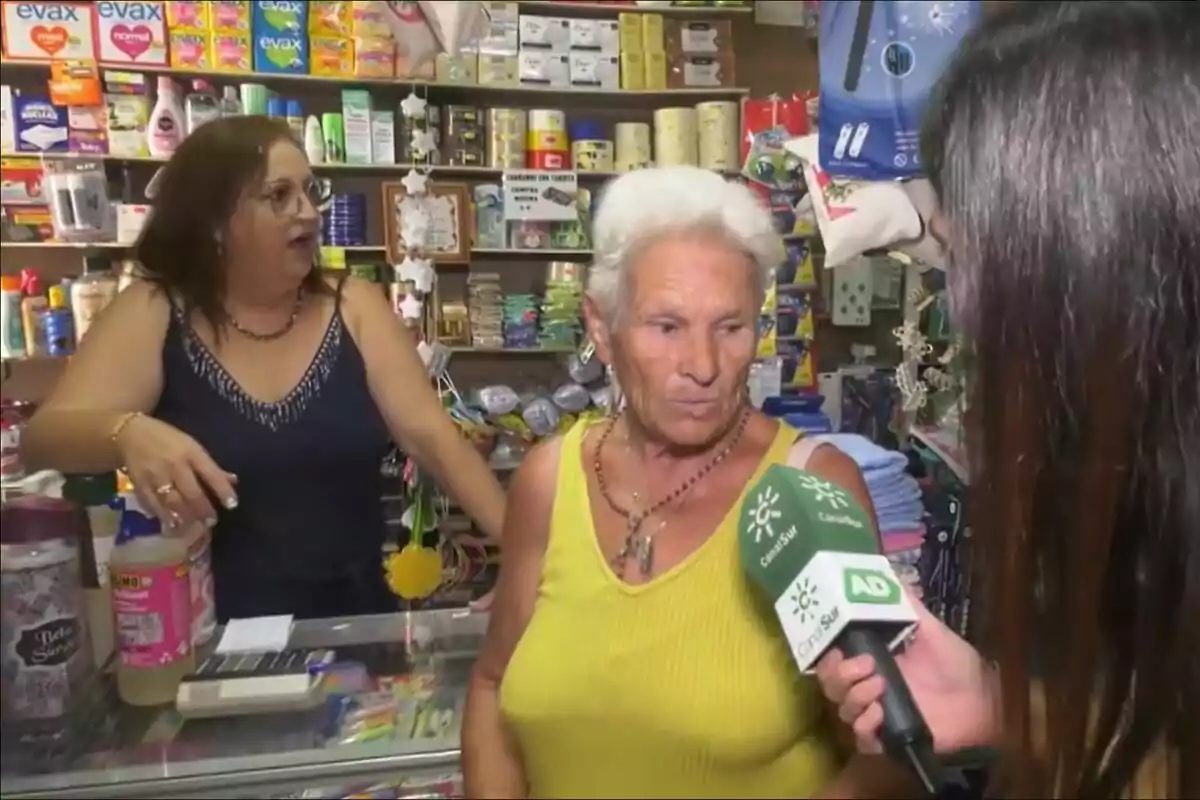 Una mujer mayor con cabello blanco y camiseta amarilla está siendo entrevistada en una tienda para Andalucía Directo en Canal Sur, mientras otra mujer con gafas y camiseta oscura observa desde el mostrador lleno de productos.