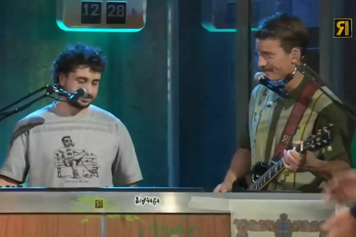 Sergio Bezos y Grison están tocando en un escenario de La Revuelta, uno toca el teclado y el otro la guitarra, ambos cantan frente a micrófonos. Sergio Bezos y Grison están tocando en un escenario de La Revuelta, uno toca el teclado y el otro la guitarra, ambos cantan frente a micrófonos.