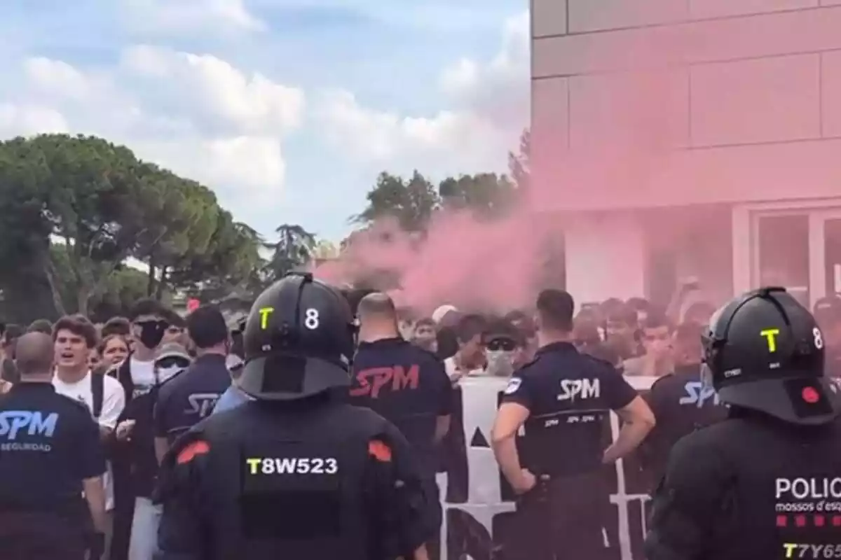 Plano general de una protesta de radicales antifascistas con un cordón policial de los Mossos en primer plano y humo rojo de fondo
