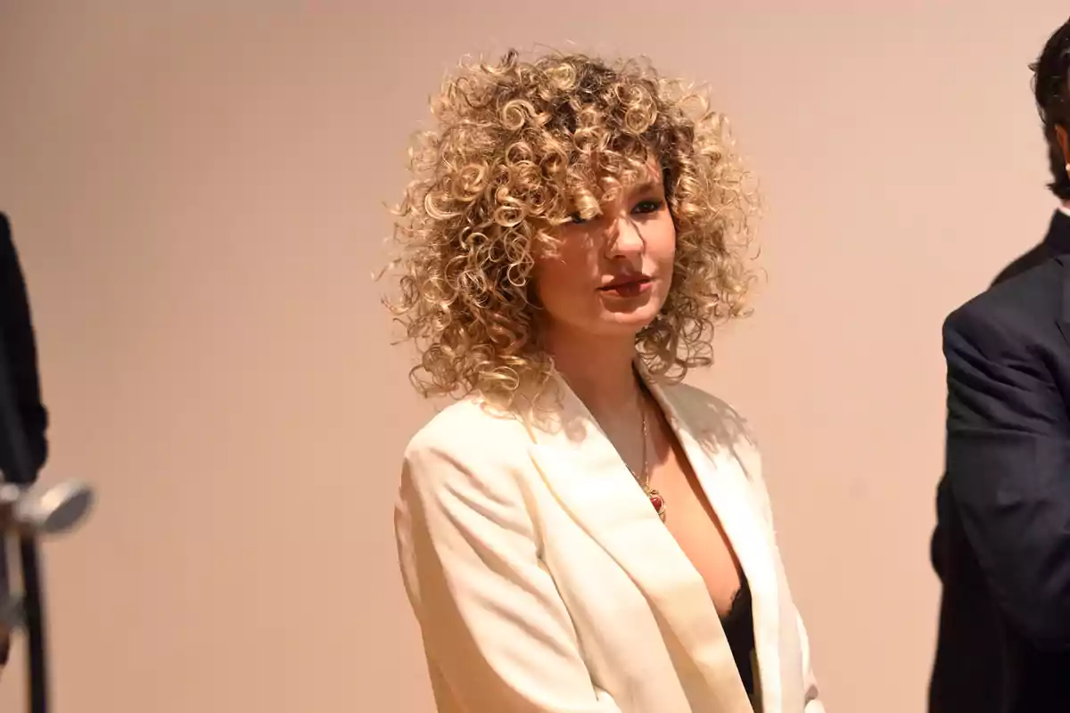 Sheila Casas con cabello rizado y rubio lleva un traje blanco en un evento formal. Sheila Casas con cabello rizado y rubio lleva un traje blanco en un evento formal.