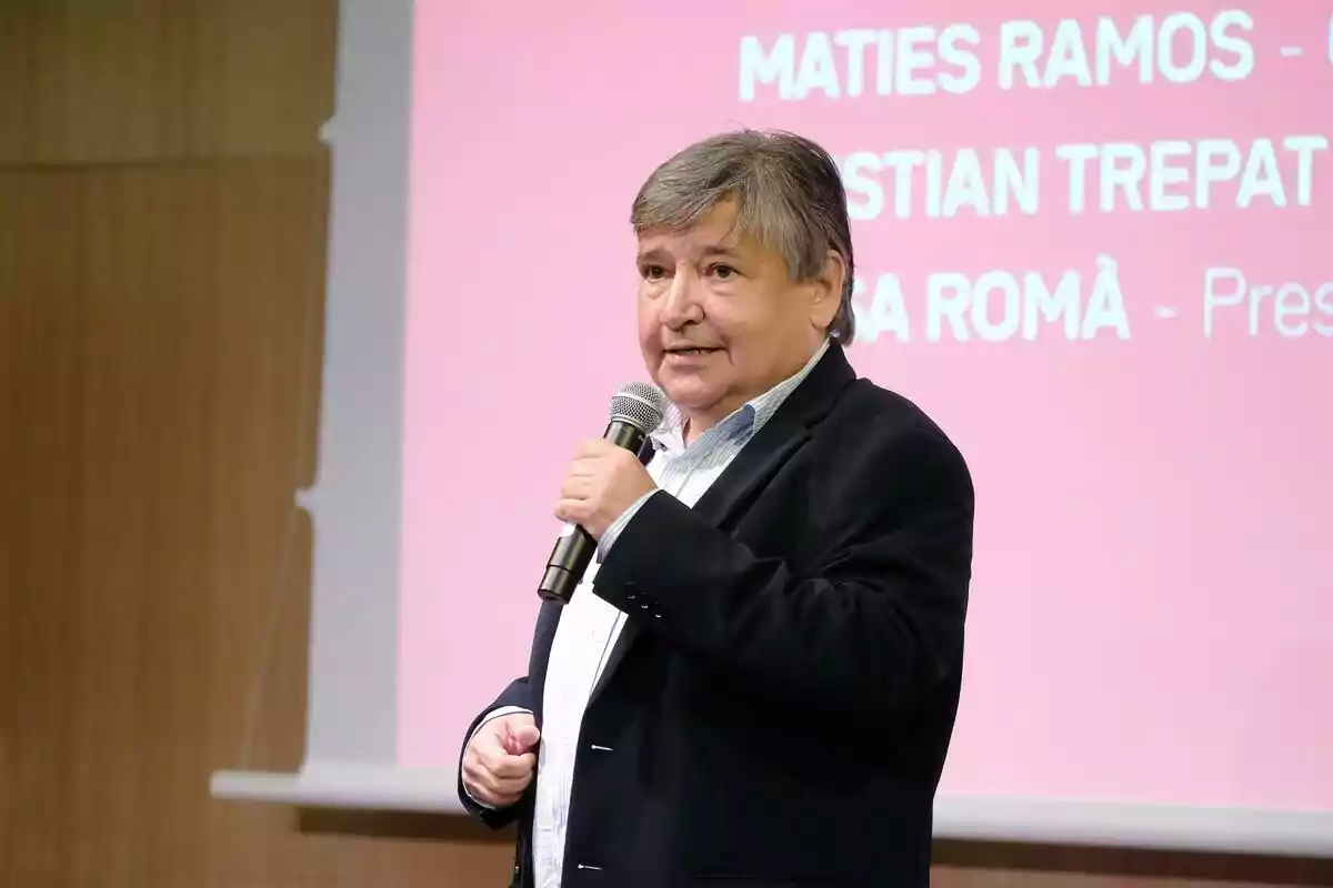 Sigfrid Gras de cabello canoso hablando por micrófono frente a una pantalla con fondo rosa y texto blanco de 3Cat. Sigfrid Gras de cabello canoso hablando por micrófono frente a una pantalla con fondo rosa y texto blanco de 3Cat.