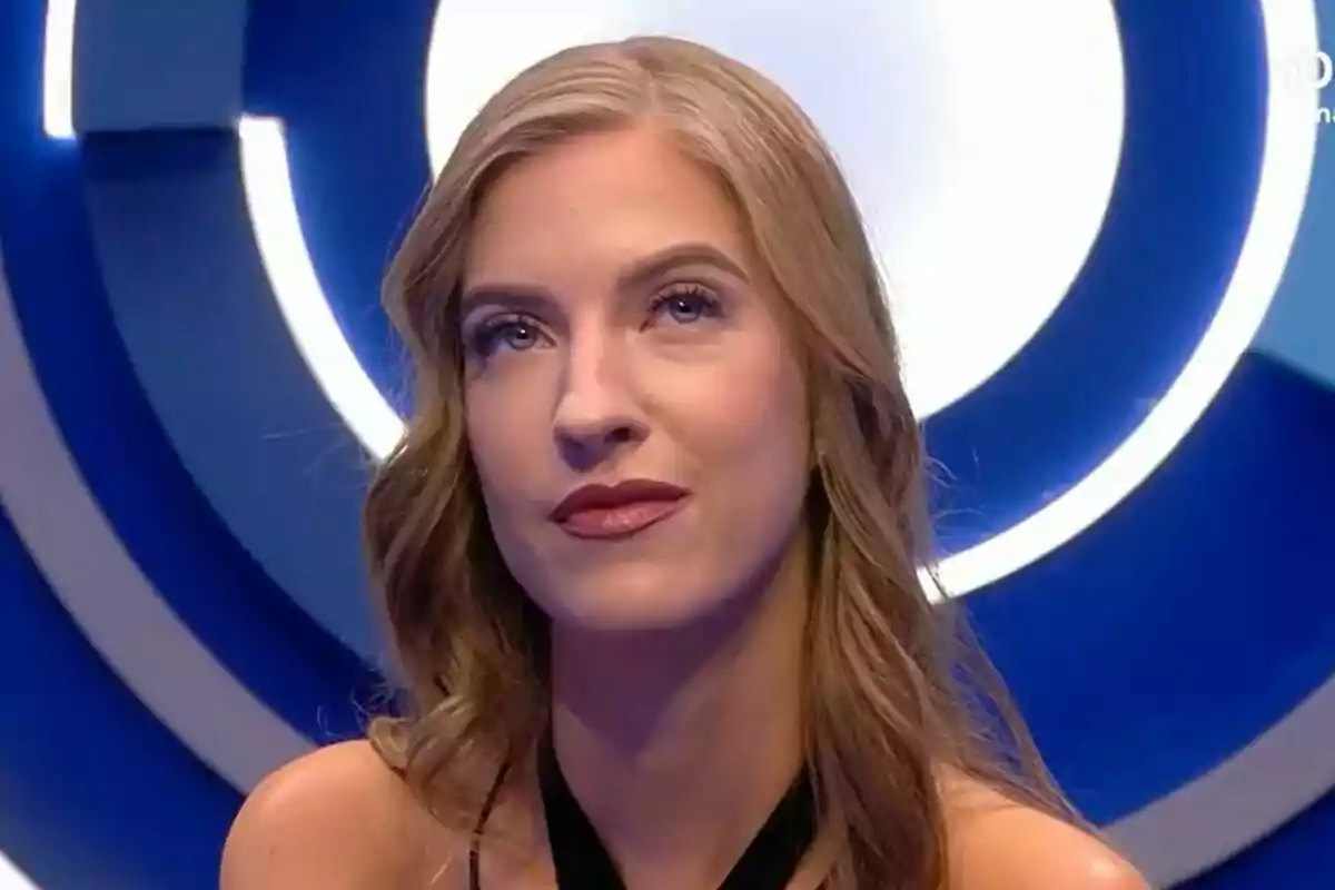 Silvia con cabello rubio y ondulado, vestida de negro, está en un estudio con luces circulares azules y blancas de fondo en el confesionario de Gran Hermano.