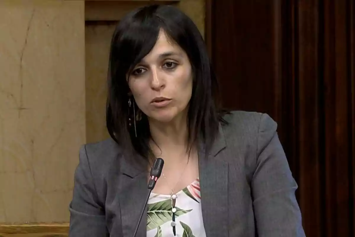 Una mujer con cabello oscuro y chaqueta gris habla frente a un micrófono en un entorno formal.