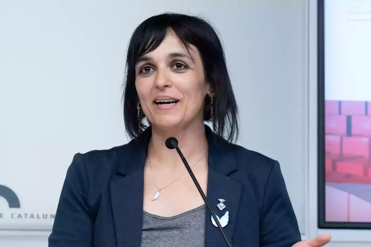 Mujer de cabello oscuro y corto hablando frente a un micrófono, vestida con una chaqueta azul y una camiseta gris, con un fondo blanco y una pantalla a la derecha.