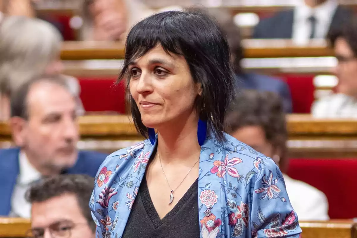 Una mujer con cabello corto y oscuro, usando una chaqueta azul con estampado floral y pendientes azules, está de pie en lo que parece ser una sala de reuniones o un parlamento, con varias personas desenfocadas en el fondo.