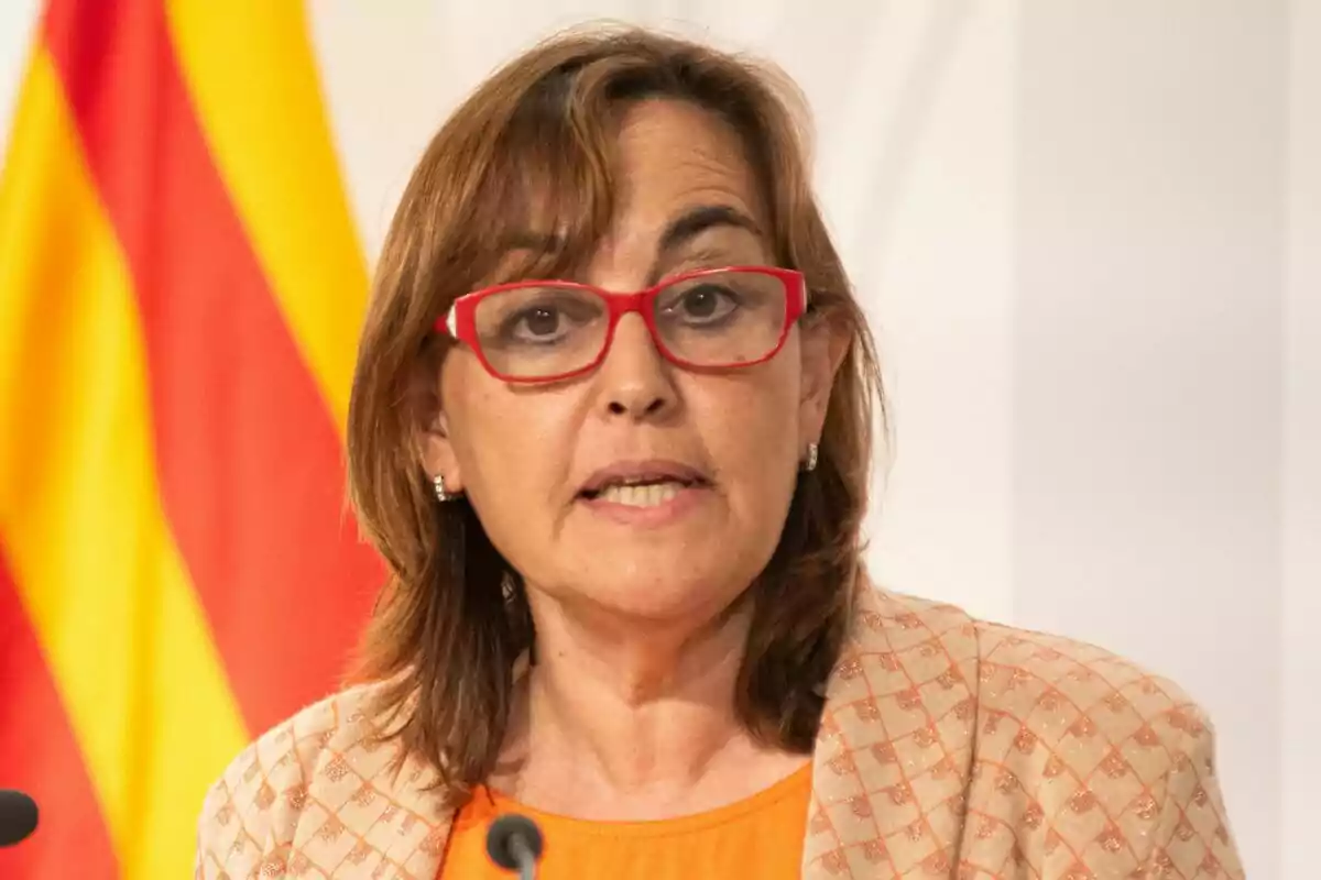 Sílvia Paneque hablando frente a un micrófono con una bandera de fondo.