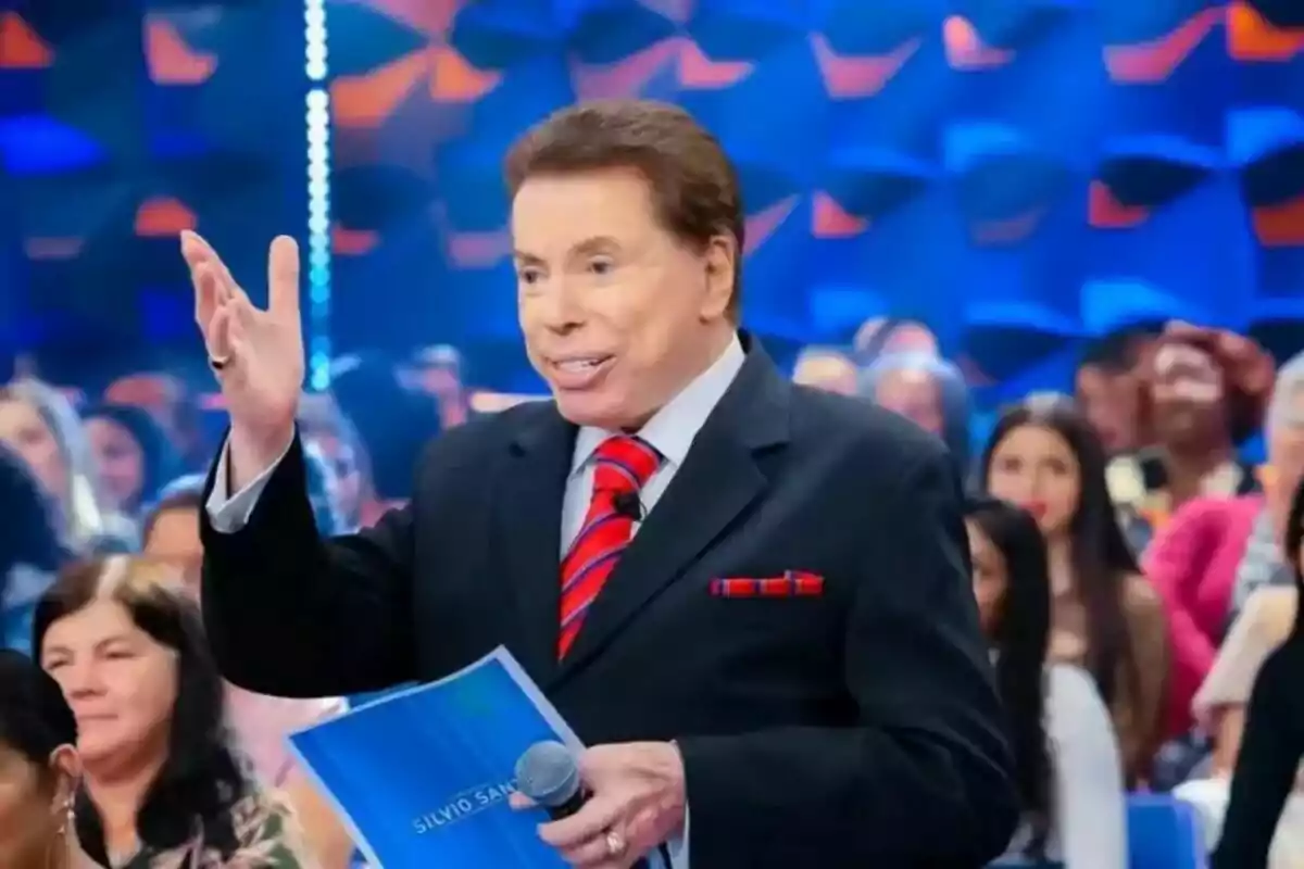 Fotografía de Silvio Santos en traje y corbata roja sosteniendo un micrófono y un documento en un escenario con una audiencia al fondo.