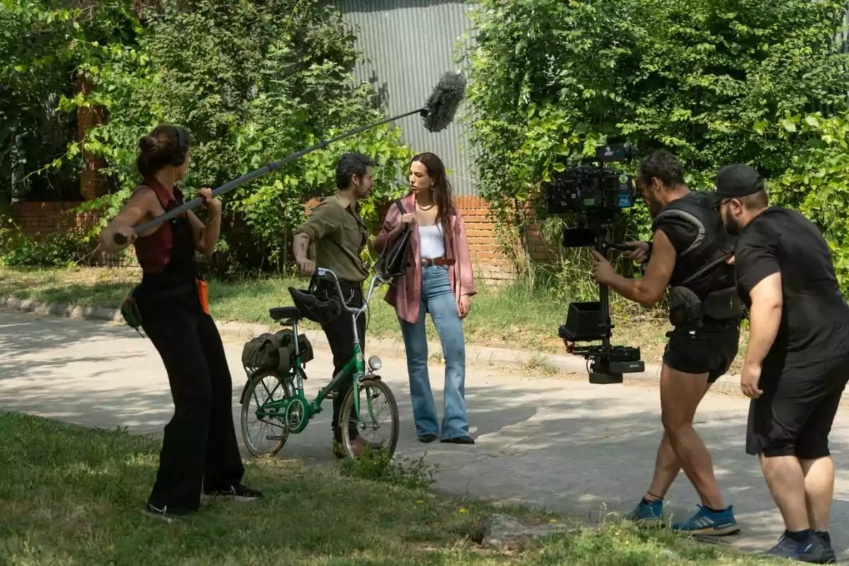 Equipo de filmación grabando una escena al aire libre donde una mujer y un hombre con bicicleta conversan mientras una persona sostiene una pértiga de micrófono y dos camarógrafos los graban en 'Sin gluten'. Equipo de filmación grabando una escena al aire libre donde una mujer y un hombre con bicicleta conversan mientras una persona sostiene una pértiga de micrófono y dos camarógrafos los graban en 'Sin gluten'.