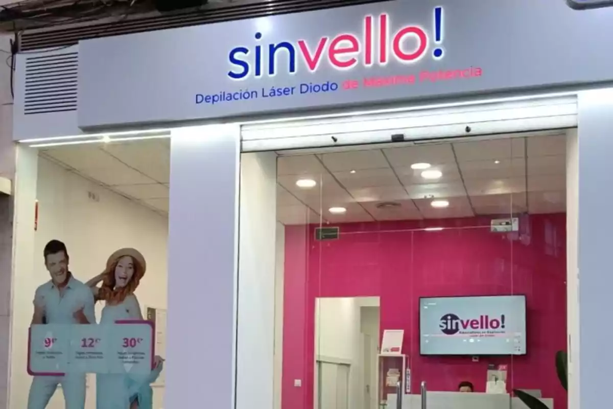 Fachada de un centro de depilación láser llamado sinvello con paredes blancas y detalles en rosa, un cartel promocional con dos personas sonrientes y una pantalla en el interior mostrando el logo del negocio