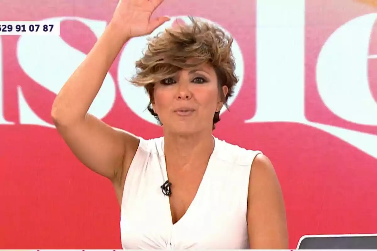 Sonsoles Ónega con cabello corto y despeinado, vestida con una blusa blanca sin mangas, levanta la mano mientras está en un set de televisión con un fondo rojo y blanco en Y ahora Sonsoles