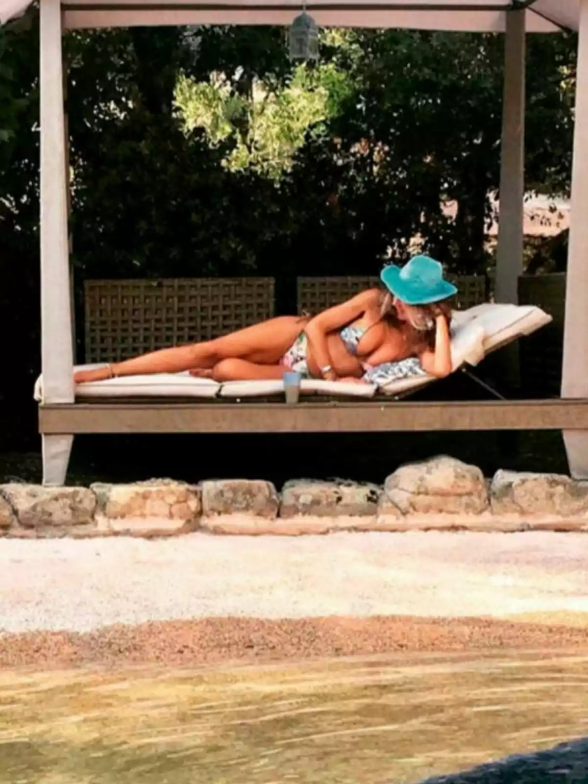 Marisa Martín-Blázquez está recostada en una cama al aire libre bajo un toldo, usando un sombrero azul y un bikini, junto a una piscina y rodeada de vegetación. Marisa Martín-Blázquez está recostada en una cama al aire libre bajo un toldo, usando un sombrero azul y un bikini, junto a una piscina y rodeada de vegetación.