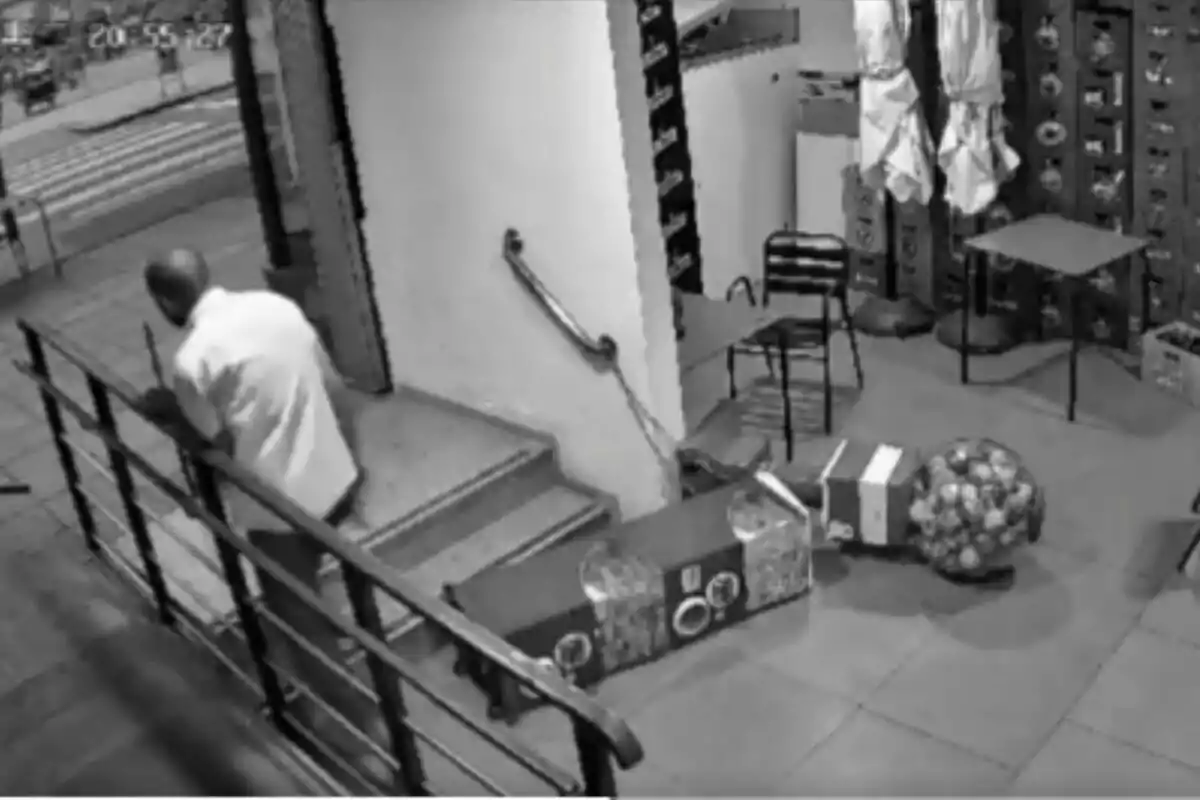 Un hombre con camisa blanca está bajando unas escaleras en lo que parece ser el interior de una tienda, donde hay varias cajas y una máquina de chicles en el suelo.