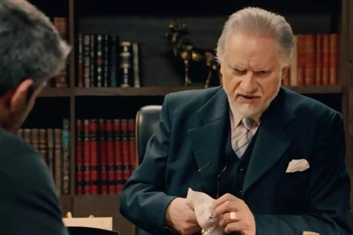 Damián está sentado en una oficina elegante con estanterías llenas de libros, lleva un traje formal y limpia sus lentes con un pañuelo mientras conversa con Andrés. Captura de 'Sueños de libertad' Damián está sentado en una oficina elegante con estanterías llenas de libros, lleva un traje formal y limpia sus lentes con un pañuelo mientras conversa con Andrés. Captura de 'Sueños de libertad'