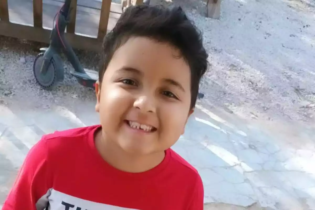 Un niño sonriente con una camiseta roja y un scooter en el fondo.