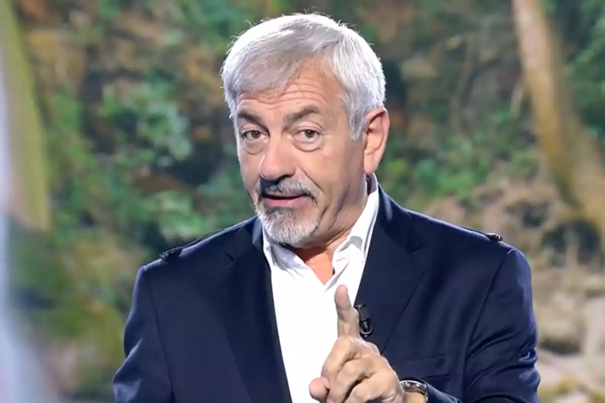 Carlos Sobera está hablando y gesticulando con el dedo índice levantado, vistiendo un traje oscuro y camisa blanca, con un fondo de naturaleza desenfocado. Captura de 'Supervivientes'