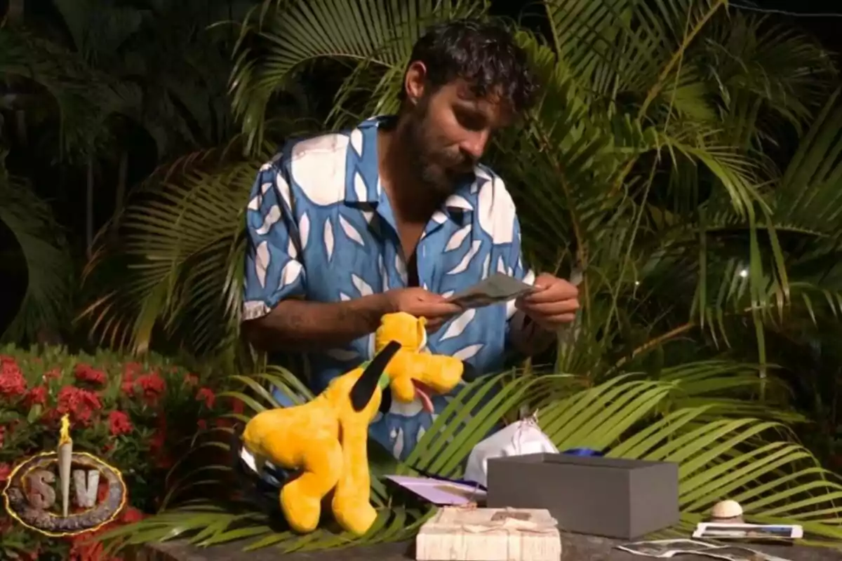 Manuel González con camisa azul y blanca observa una foto mientras está rodeado de vegetación y objetos como un peluche amarillo y una caja gris. Captura de 'Supervivientes' Manuel González con camisa azul y blanca observa una foto mientras está rodeado de vegetación y objetos como un peluche amarillo y una caja gris. Captura de 'Supervivientes'