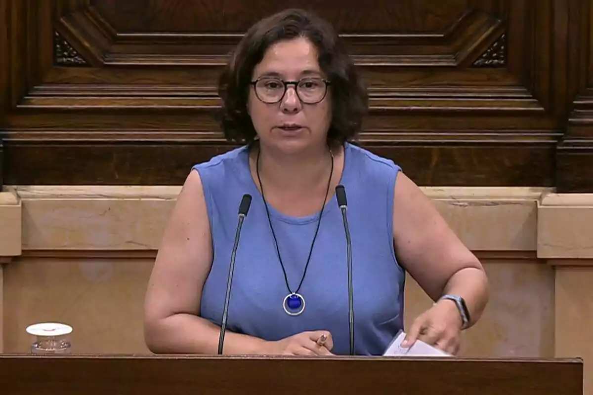 Mujer de cabello corto y gafas hablando en un atril con dos micrófonos en un entorno institucional Mujer de cabello corto y gafas hablando en un atril con dos micrófonos en un entorno institucional