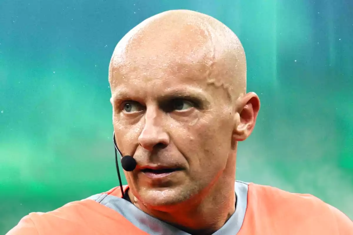 Árbitro calvo con auricular y camiseta naranja en un estadio con fondo verde difuminado