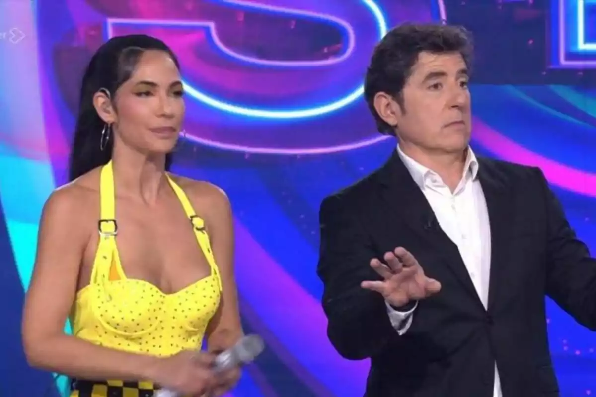 Dos personas en un escenario con fondo colorido de Tu Cara Me Suena, Tamara Gorro con vestido amarillo y Manel Fuentes con traje oscuro.
