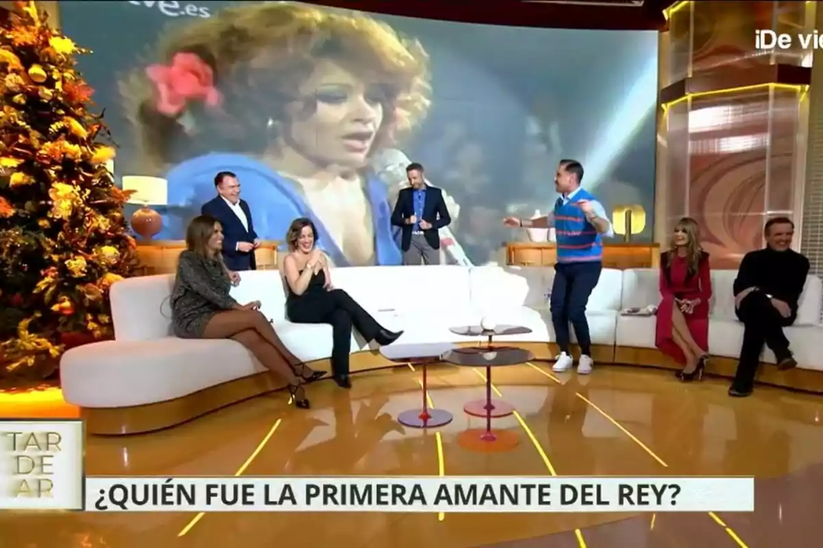 Un grupo de personas como Juan Luis Galiacho, Frank Blanco, Marta López, Leticia Requejo, Miguel Ángel Nicolás, Marisa Martín Blázquez y Antonio Montero está sentado en el plató de TardeAR decorado con un árbol de Navidad, mientras en la pantalla se proyecta la imagen de una mujer cantando.