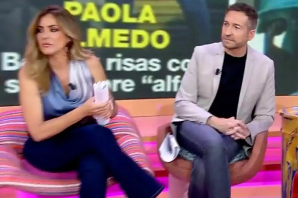 Frank Blanco y Verónica Dulanto sentados en 'Tardear' colorido con un fondo que muestra letras amarillas y blancas Frank Blanco y Verónica Dulanto sentados en 'Tardear' colorido con un fondo que muestra letras amarillas y blancas