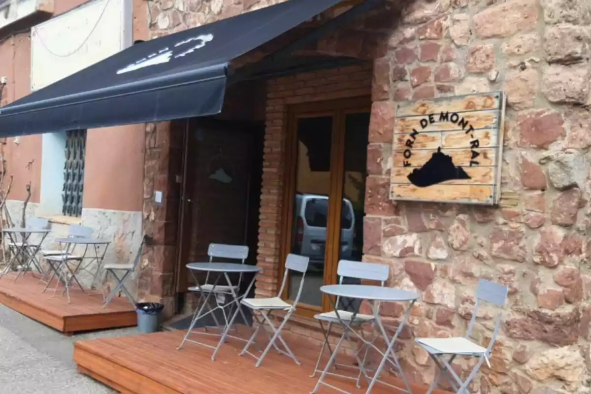 Fachada de cafetería con mesas y sillas metálicas en una terraza de madera junto a una pared de piedra y un letrero de madera que dice Forma de Mont-ral Fachada de cafetería con mesas y sillas metálicas en una terraza de madera junto a una pared de piedra y un letrero de madera que dice Forma de Mont-ral