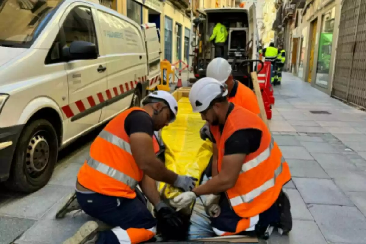 Trabajadores de la construcción con chalecos naranjas y cascos blancos realizan tareas en una calle junto a una furgoneta blanca.