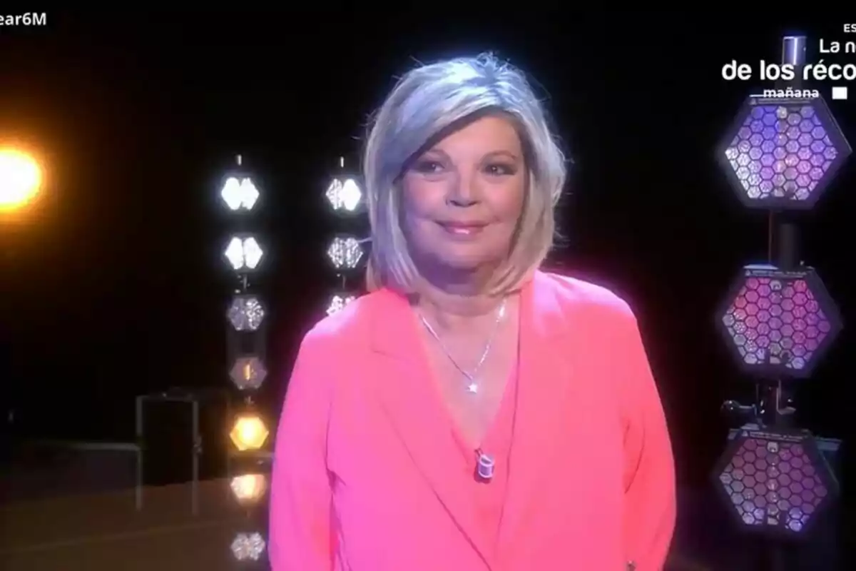 Terelu Campos con cabello rubio y un atuendo rosa está de pie frente a un fondo oscuro con luces hexagonales en Tardear. Terelu Campos con cabello rubio y un atuendo rosa está de pie frente a un fondo oscuro con luces hexagonales en Tardear.