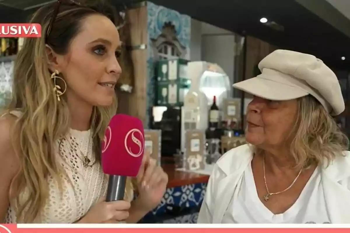 Una reportera sostiene un micrófono rosa de Socialité mientras entrevista a Terelu Campos de cabello rubio y gorra beige en un lugar cerrado. Una reportera sostiene un micrófono rosa de Socialité mientras entrevista a Terelu Campos de cabello rubio y gorra beige en un lugar cerrado.