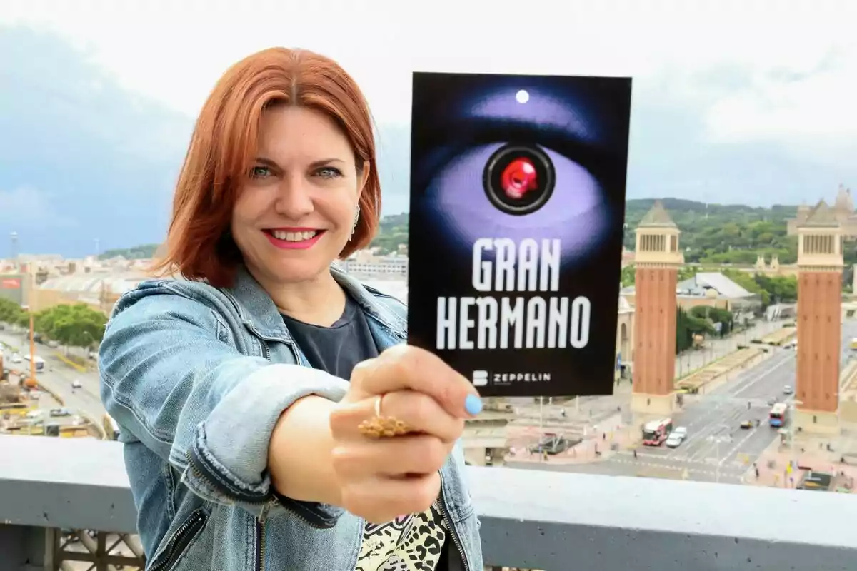 Fotografía de Teresa Colomina sonriente con cabello rojo sostiene un cartel del programa "Gran Hermano" con Barcelona de fondo.