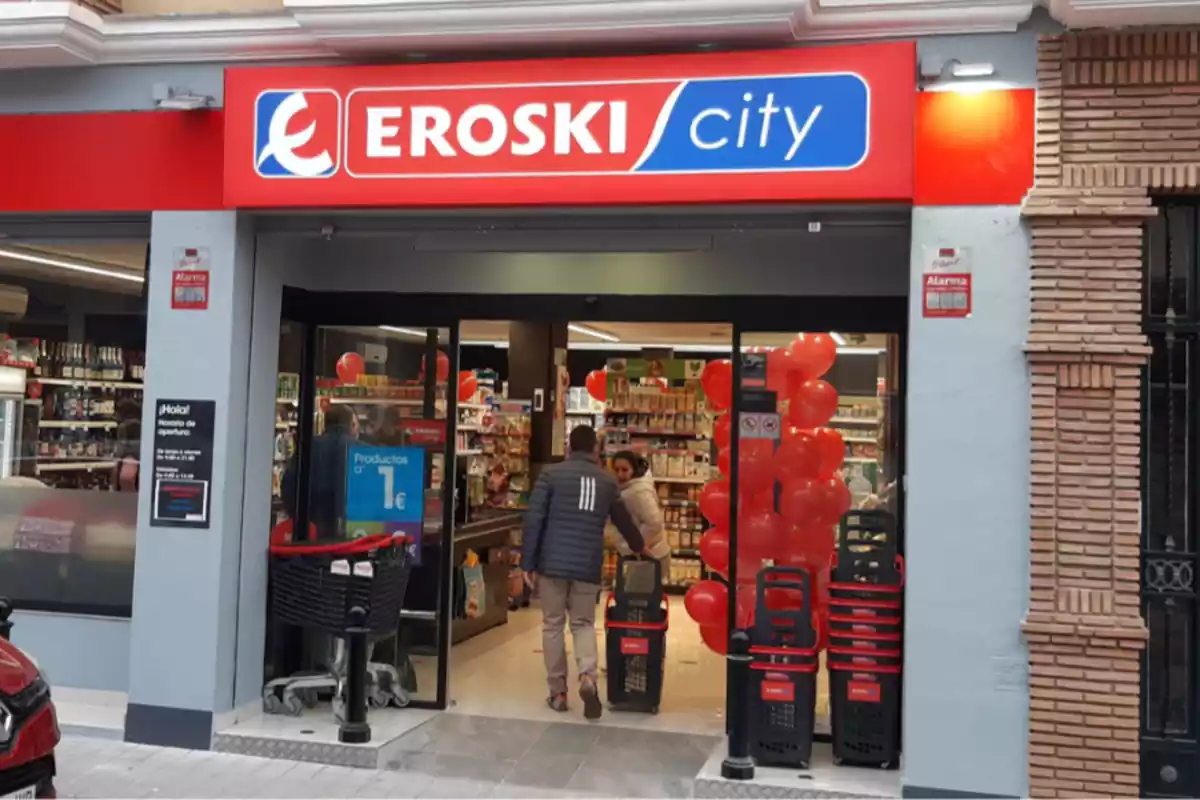 Exterior de un supermercado de Eroski y un cliente entrando por la puerta de la tienda con un carrito en la mano